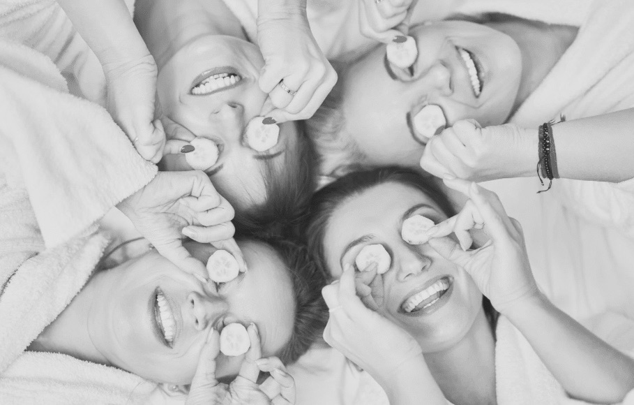 Four women laying with heads, touching in a circle, holding cucumbers to their eyes, indicating a spa ritual